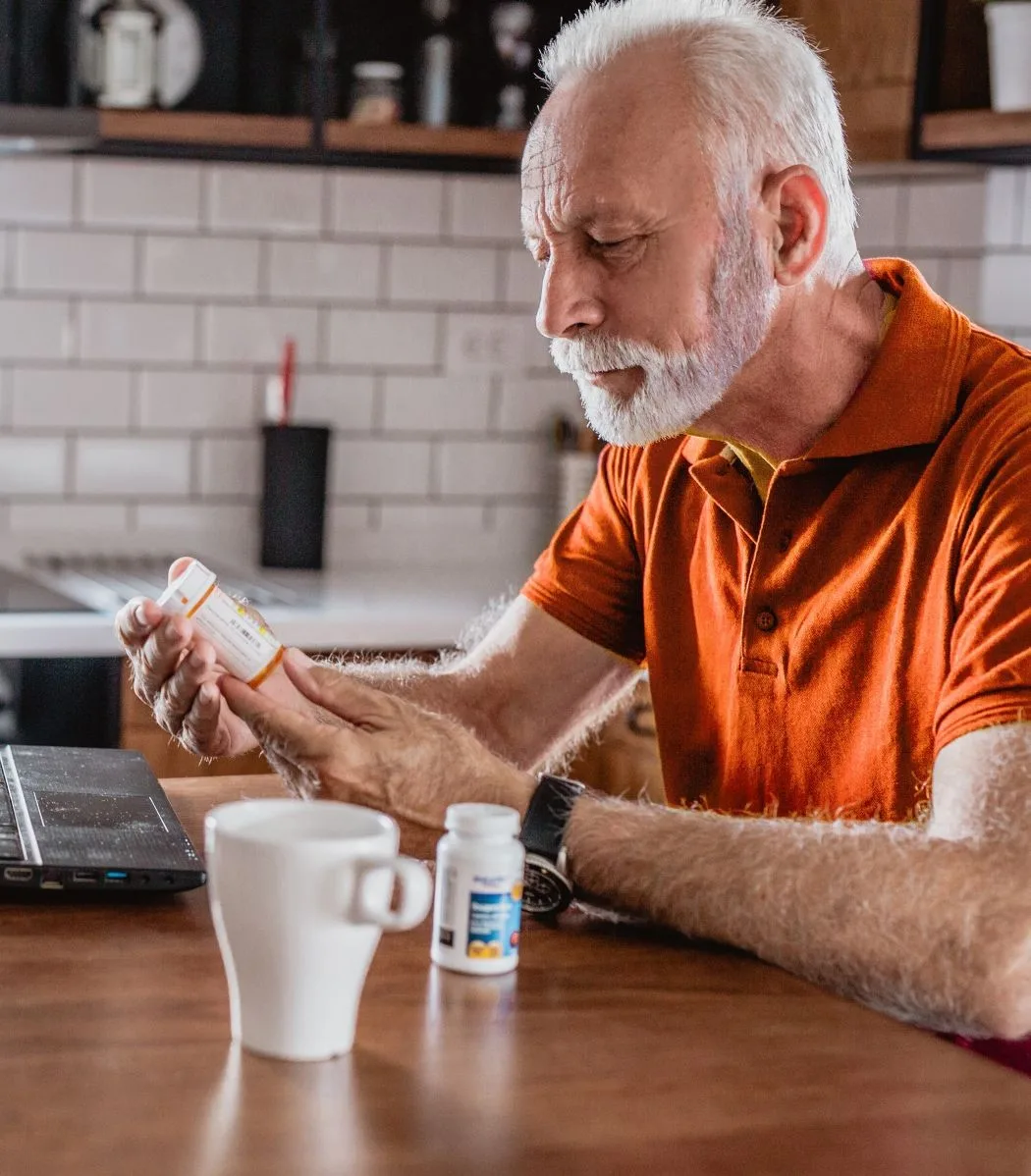 Man with white hair and beard sitting at table looking at medicine bottle